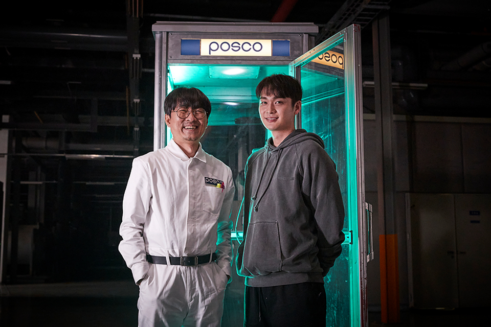 포스코 라고 쓰여진 공중전화 박스 앞에서 미소짓고 있는 장항준 감독과 조윤우 배우의 모습.