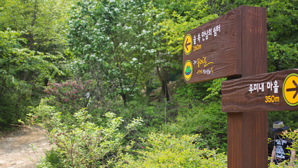 고구려 대장간 마을 아차산 둘레길 입구 표지판 모습. 나무 표지판에 오른쪽 방향의 화살표와 함께 우미내 마을 350m라고 적혀있고, 왼쪽 방향의 화살표화 함께 숲 속 만남의 쉼터 260m라고 적혀있다.