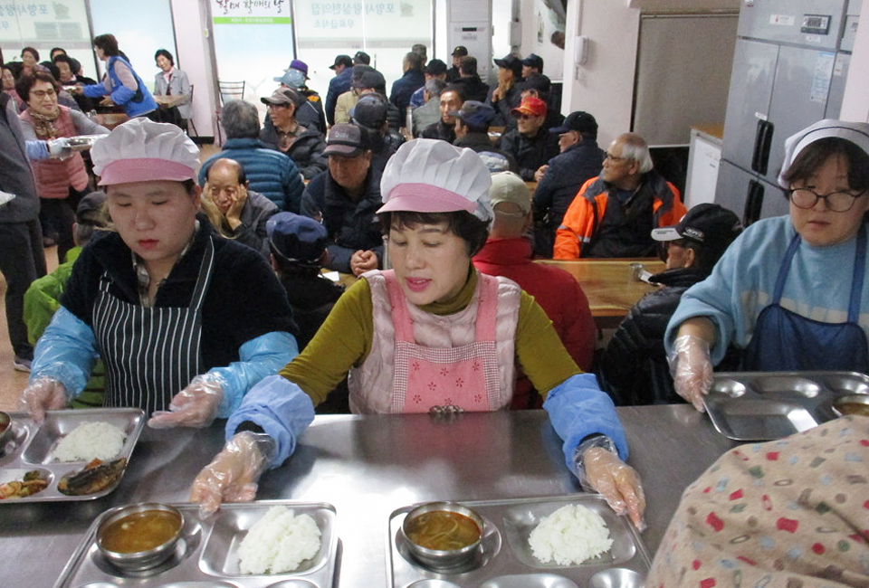 급식소의 모습. 뒤로 급식소를 이용하는 어르신들의 모습과 가운데 위상모자와 위생장갑을 착용하고 배식하는 세 사람의 모습.