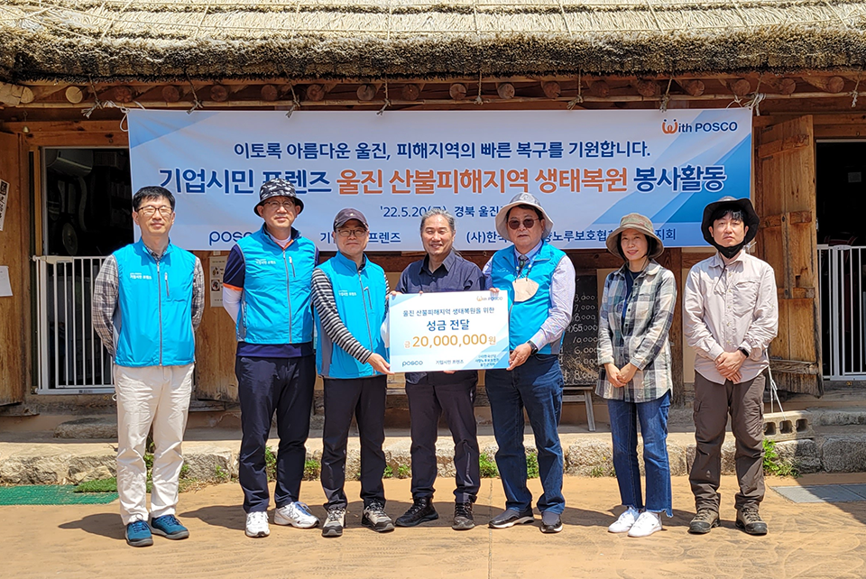 '이토록 아름다운 울진, 피해지역의 빠른 복구를 기원합니다. 기업시민 프렌즈 울진 산불피해지역 생태복원 봉사활동'이라고 적힌 현수막을 배경삼아 하늘색 조끼를 입은 남자 네명과 체크무늬옷 여자 한명 하얀 셔츠 한명 남색셔츠 한명이 나란히 서있으며, '울진 산불피해지역 생태복원을 위한 성금 전달 금 20,000,000원이라고 적힌 팻말을 들고있다.