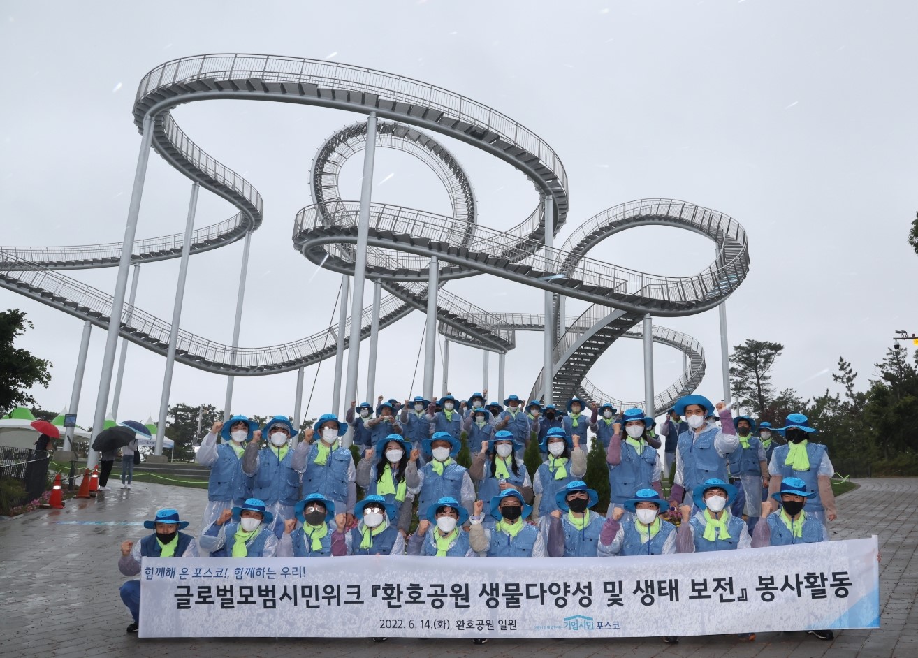 하늘색 모자와 조끼 그리고 우비를 입고 있는 봉사단과 사진 두번째 줄 왼쪽 다섯번째 김학동 부회장과 포항 스페이스워크 앞에서 "함께해 온 포스코! 함께하는 우리! 글로벌모범시민위크 [환호공원 생물다양성 및 생태 보전] 봉사활동이라고 적힌 현수막을 들고 화이팅 하며 사진을 찍고있다.