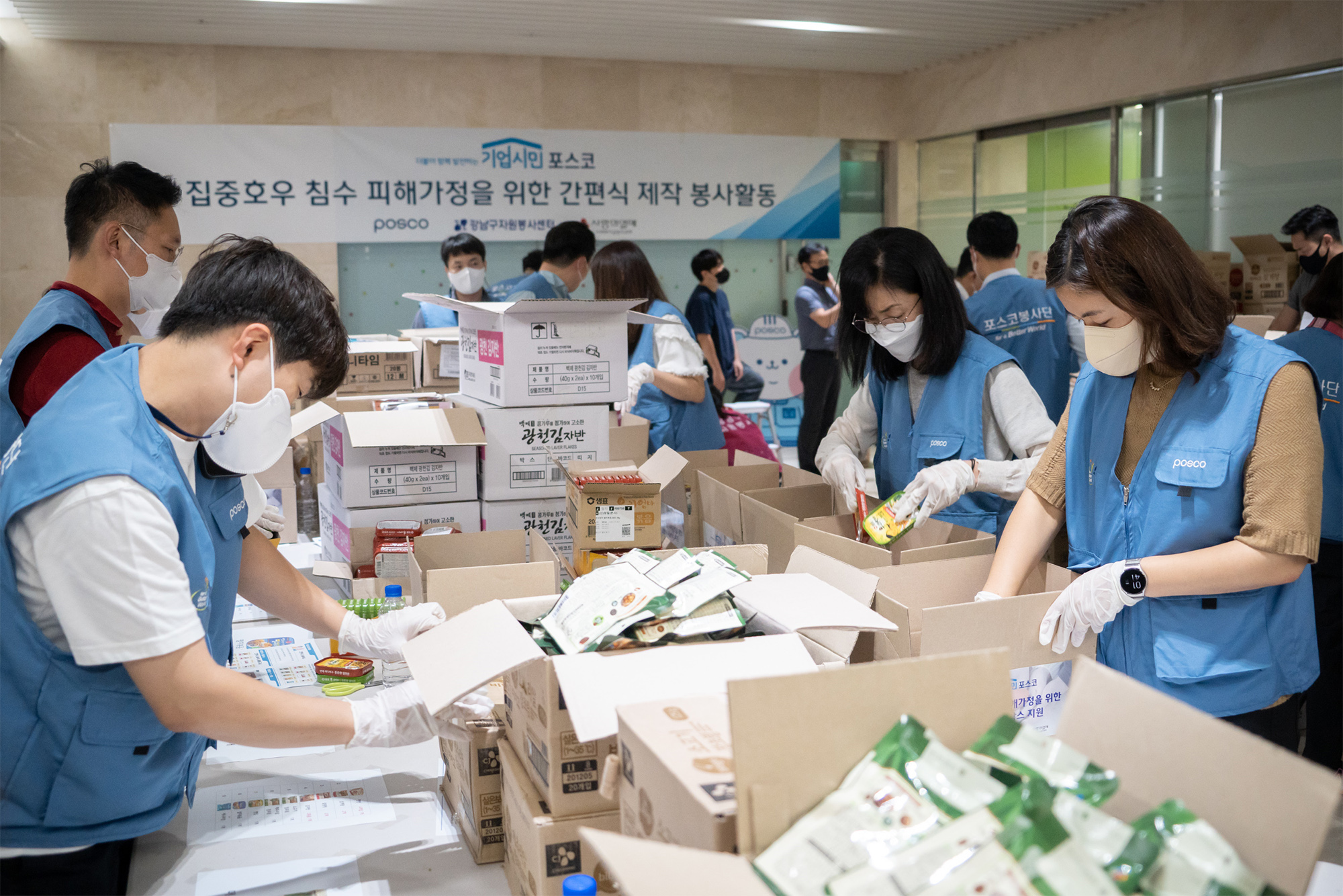 여러명의 사람들이 테이블 위에 쌓여있는 박스에 물건들을 정리하고 있는 모습이다.