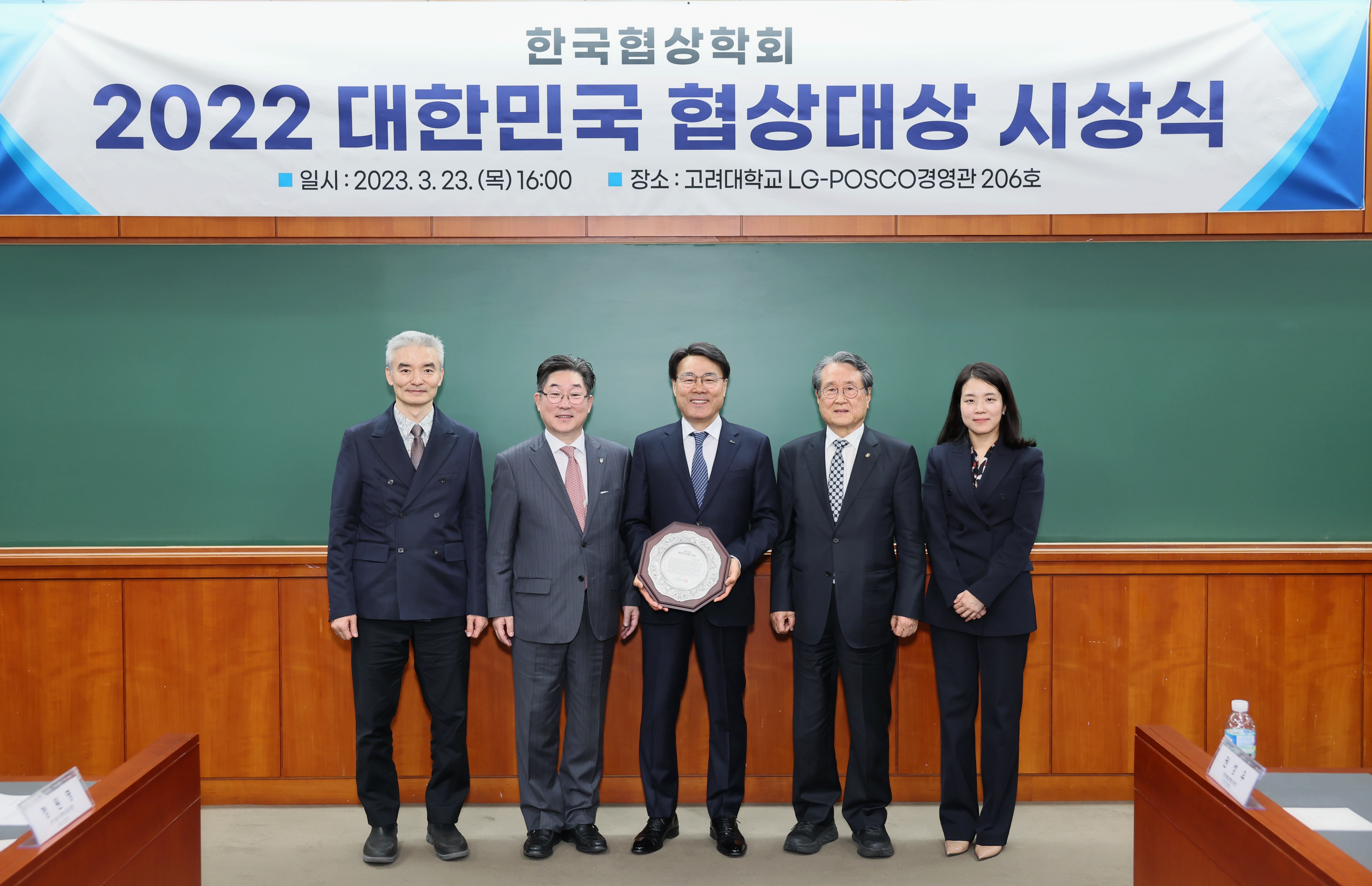 한국협상학회 2022 대한민국 협상대상 시상식 이라는 현수막 앞에서 정장 입은 남자 4명, 여자 1명이 나란히 서있으며, 가운데 남성 한명은 팔각형 상패를 두손에 들고 기념촬영하고 있는 모습이다.