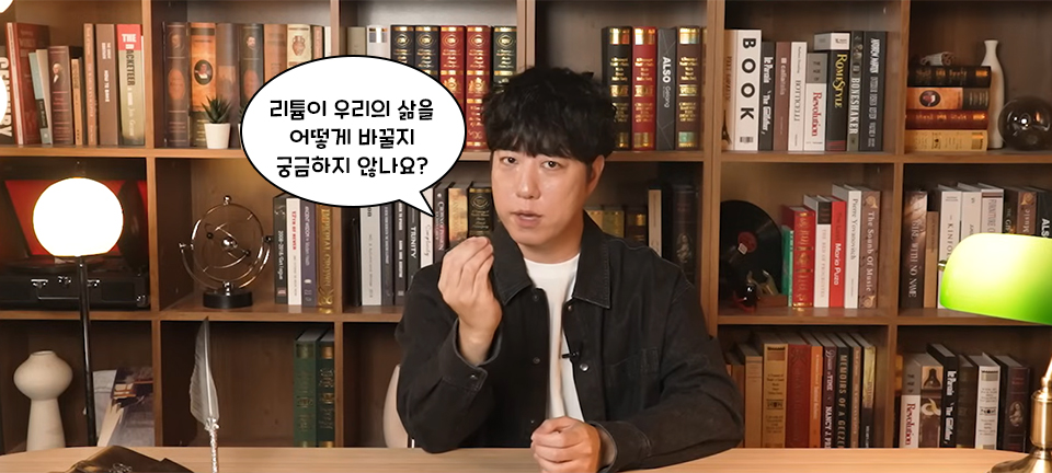 "리튬이 우리의 삶을 어떻게 바꿀지 궁금하지 않나요? " 라고 질문하며 한 손을 들어보이는 서재 책상 앞에 앉아 카메라를 보는 궤도의 사진. 