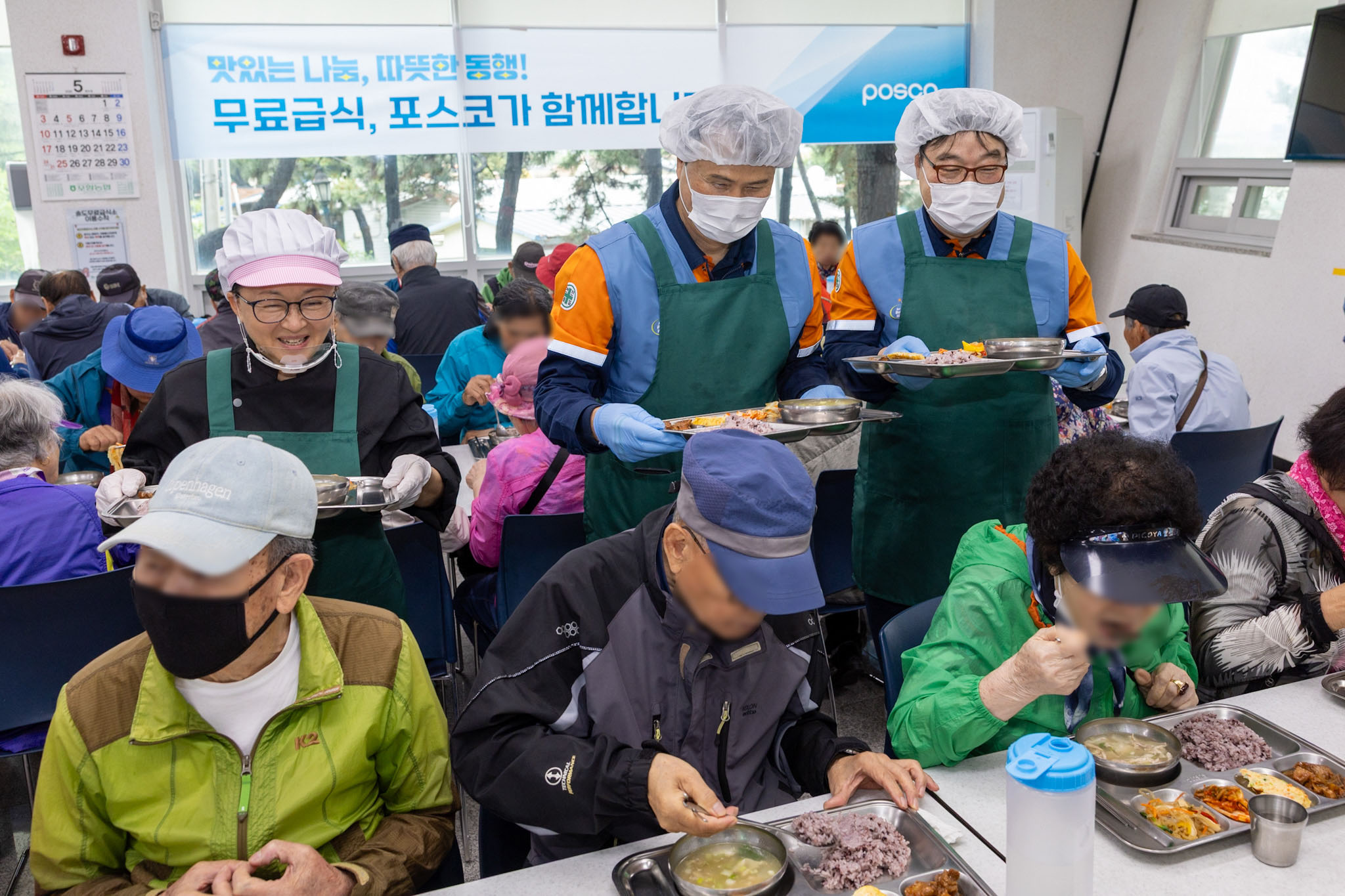 이희근 포스코 사장(가운데)과 급식대가 이미영 셰프, 오창석 포항제철소 제강부 명장이 마스크와 위생 모자를 쓰고, 식판을 들고 식사하고 있는 어르신들 사이에서 환하게 웃고 있다.
