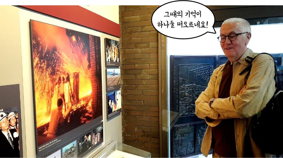 포스코역사박물관에서 과거 제철소 작업 사진을 관람하고 있는 프랑스 엔지니어 제라르 르벨 님의 모습이다. 제라르 르벨 님은 팔짱을 낀 채 미소를 지으며 전시를 바라보고 있으며, 머리 위에는 '그때의 기억이 하나둘 떠오르네요!'라는 문구가 적힌 말풍선이 있다. 왼쪽 벽면에는 붉은 쇳물이 강렬하게 튀는 용광로 작업 현장 사진이 크게 걸려 있다.
