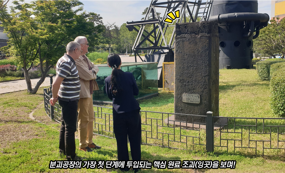 야외 전시장에서 두 명의 외국인 방문객과 여성 가이드가 '조괴(잉곳)' 전시물을 바라보고 있는 모습이다. 전시물은 검고 거친 표면의 직사각형 기둥 모양 쇳덩어리이며, 윗부분에는 노란색 강조 표시 아이콘이 그려져 있다. 하단에는 '분괴공장의 가장 첫 단계에 투입되는 핵심 원료 조괴(잉곳)를 보며!'라는 자막이 적혀 있다.